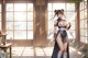 A woman in traditional attire stands indoors with sunlight streaming through windows.